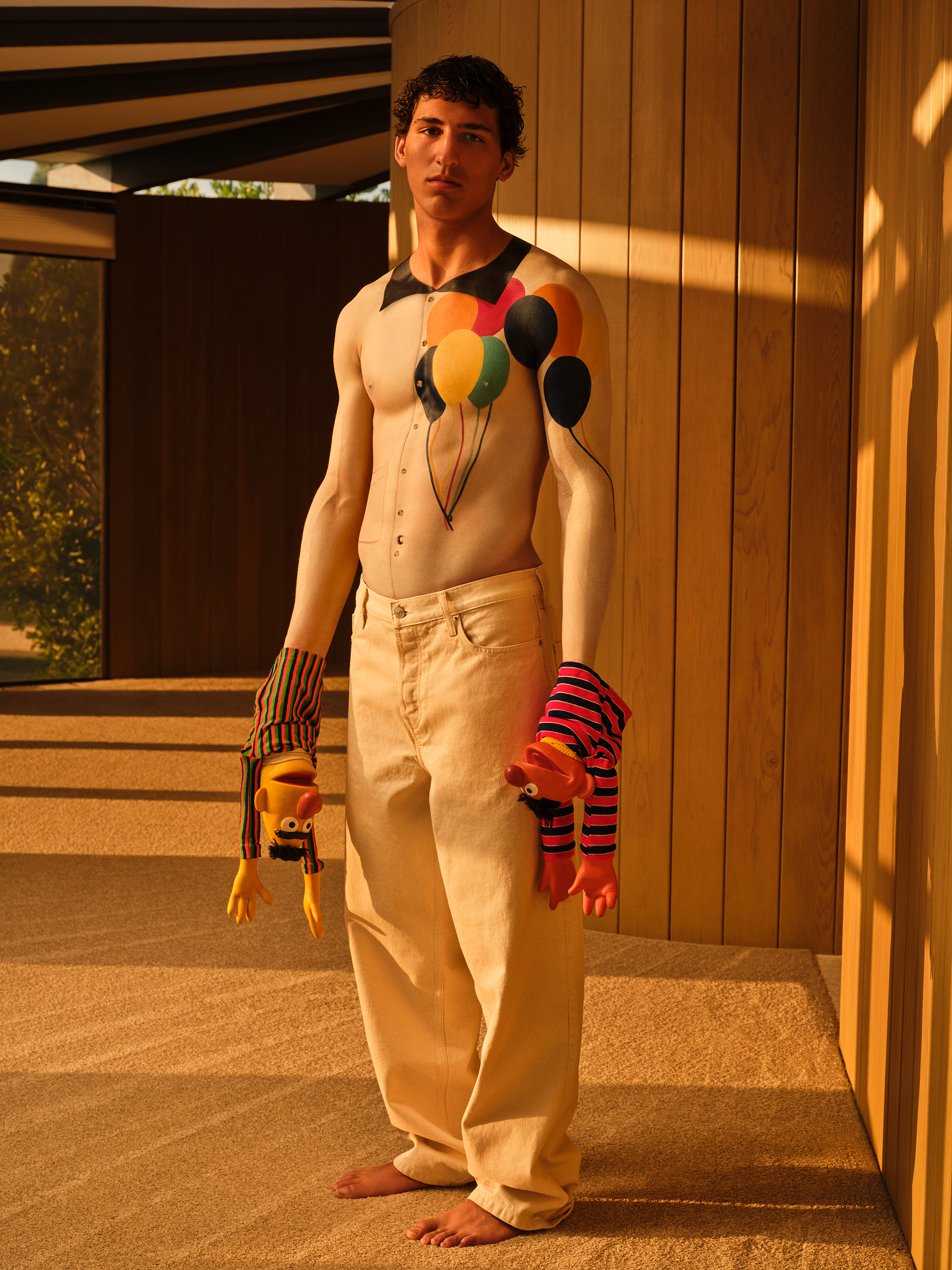 Male model with a white body painted top with baloon graphics holding bert and ernie puppets and looking at the camera in a warmly lit room